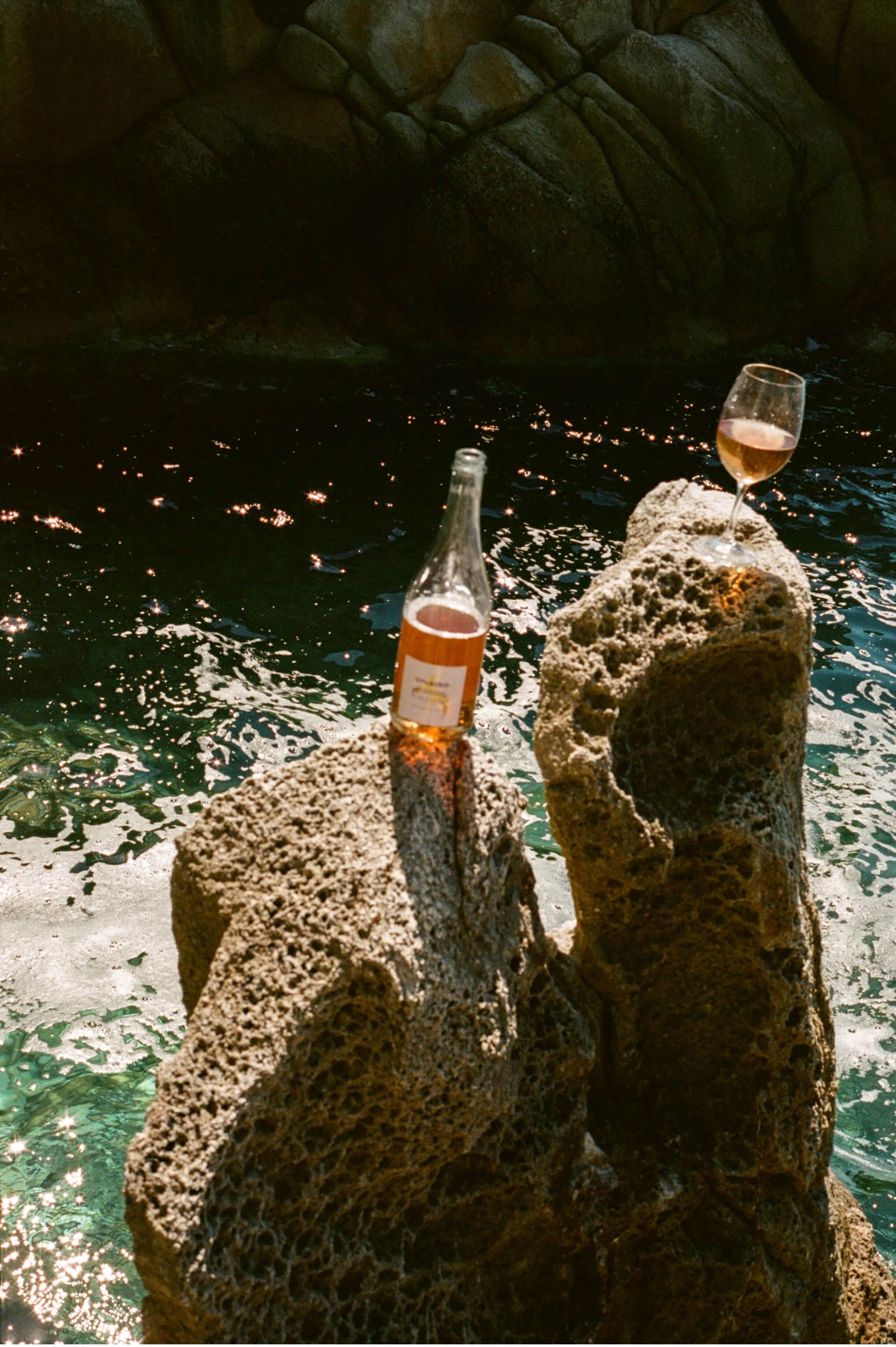 Bottle and glass of orange wine on a rock with water in the background